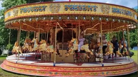 States of Guernsey Pride of the South carousel. It is predominately red and gold with children riding carousel horses. The photo has been taken on a sunny but cloudy day.