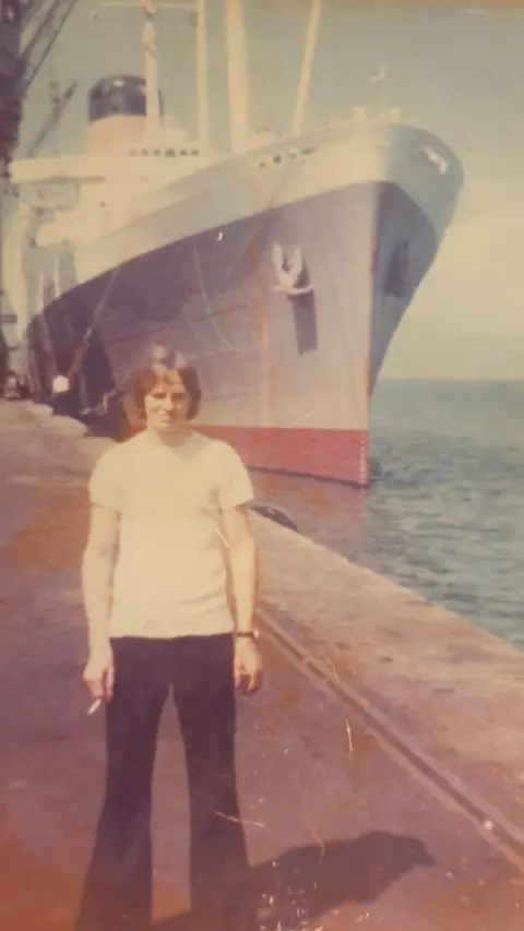 Helen Clark A faded colour image from the 1970s shows Tommy Jones in the foreground wearing dark bell bottom jeans and a white T-shirt. He has a wide leg stance, is holding a cigarette in his right hand and has light brown hair in a pageboy-style cut. He is standing near the edge of a dock, dwarfed by a black and white cruise liner behind him. The red paint from below the ship's waterline is visible as is its bow anchor and a funnel.