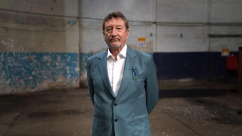 Peaky Blinders writer Steven Knight, wearing a turquoise suit jacket and white shirt, standing in an empty warehouse-style room at Digbeth Studios