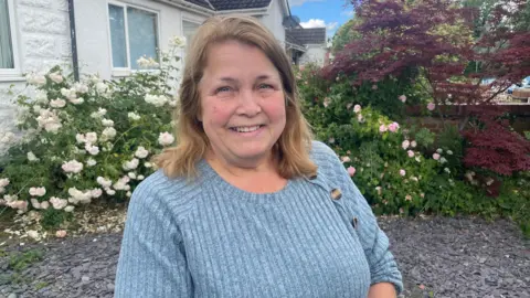 BBC A woman with shoulder-length fair hair, smiling, wearing a ribbed grey-blue jumper. She stands in a garden in front of climbing pink and white roses. A white house, a red-leaved tree and a bright sky with light cloud sit behind her.