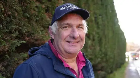 Shaun Whitmore/BBC Steve Nott stands in front of a large green hedge and smiles at the camera. He wears a navy cap, navy jacket and red polo top. 