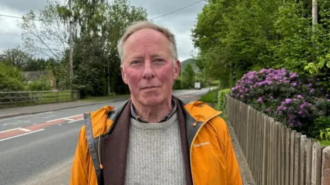 Charlie Stubbs / BBC Chris is wearing an orange Montane coat with a brown lining, a scarf and a grey jumper. He has grey short hair, which is pushed to one side. He is stood in front of the A49. There is a wooden fence on the right of the picture, with big trees and a purple plant. 