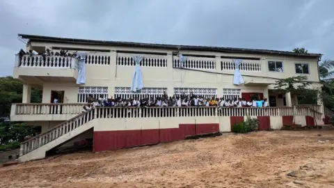 Go The Extra Mile A two‑storey cream‑coloured building with red paint on the lower walls and decorative railings. A large group students, stand on the upper balcony. The area in front of the school is sandy. Curtains hang from the upper windows, and small plants and trees surround the building. The sky is overcast.