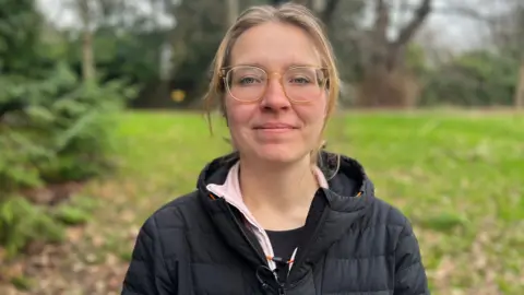 BBC Karolina Pakenaite. She has long blonde hair which is tied up and glasses. She is wearing a black puffer coat over a pink fleece and a black t-shirt. She is pictured outside, standing in front of a field and some trees. She is looking directly at the camera and smiling. 