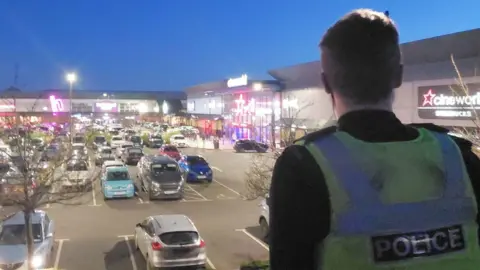 A police officer stands with his back to us. He is watching over the car park at a retail park, late at night.