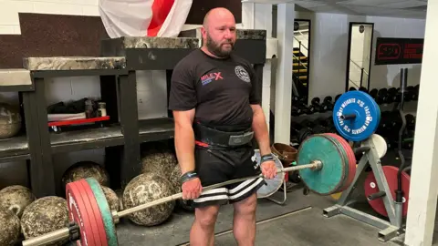 James Grant/BBC A man in a black T-shirt and shorts lifts a metal bar with lots of weights on.