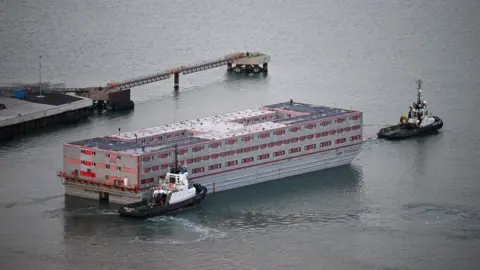 Closer image of the Bibby Stockholm as it leaves Portland. There is a tugboat at the front of it and a second tugboat to the back of it. The Bibby Stockholm is a red and white rectangular barge, moving through the water and there is a dock platform seen to the left of the image.