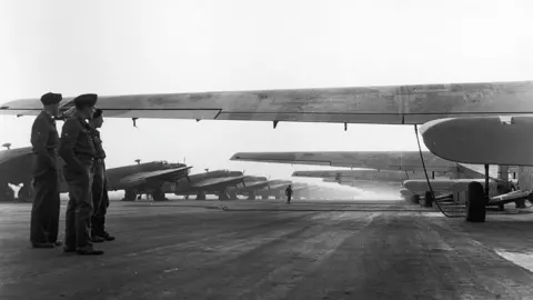 Getty Images Sebuah jalan dari Hamilcar Gliders (kanan) dan RAF Halifax Towing Aircraft (kiri) pada 24 Maret 1945. Gambar hitam dan putih menunjukkan tiga pesawat terbang dengan tangan mereka di saku mereka memandang ke arah glider.