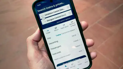 Hand holding a smartphone displaying the ScotRail app journey planner. The screen shows a search for train travel from Exhibition Centre (Glasgow) to Glasgow Central, with options for ticket type, travel date, departure time, passengers and railcards. The background is an out-of-focus tiled pavement.