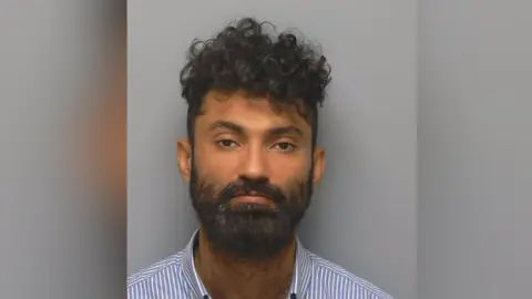 Mugshot of Diwan Khan, who is wearing a striped blue and shirt shirt and has a black beard and black curly hair.
