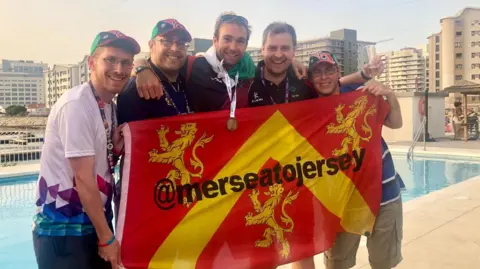 The brothers and friend, Dave, standing in front of an Anglesey county flag in front of a swimming pool