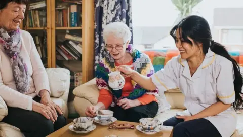A care worker pours tea for two older adults seated on a sofa in a living room, with cups and saucers arranged on a coffee table in front of them. A nurse with long dark hair is lifting a china jug. There is a bookshelf in the background.