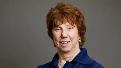 Baroness Catherine Ashton Baroness Catherine Ashton wearing a navy blue shirt smiling at the camera. She has short wavy auburn hair and is wearing droplet earrings