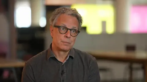 Ian Russell pictured during an interview with the BBC. He wears a grey shirt and black, round glasses.