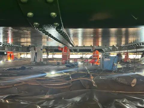 Workers in orange high-vis attire are working under a section of the Tyne Bridge. Various cables are strewn on the ground.