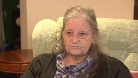 Lynda Wilkins is sitting in an armchair in the communal lounge of Kings Gardens retirement flats. She has long grey hair and is wearing a purple patterned top.