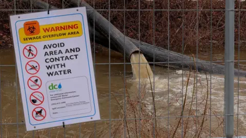 Pipes divert raw sewage away from the Potomac River. A sign on a fence warns people not to make contact with the water