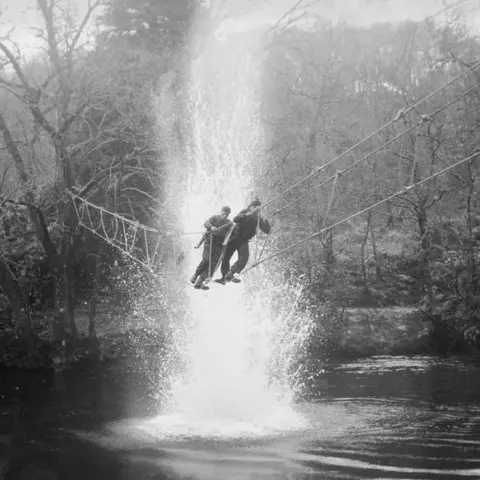 Getty Images Two commandos carrying rifles cross a rope bridge over water at Achnacarry. A plume of rises up from a small explosion in the water. Trees fringe the water's edge.