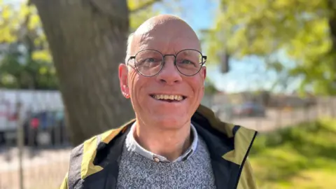 Head and shoulder image of Tim Bick. He has rounded glasses and is wearing a yellow coat, open, with a grey jumper and light shirt. He is smiling at the camera and standing in front of a tree and patch of grass. 