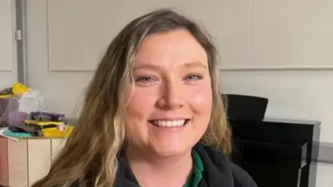 Sinéad is smiling at the camera and has long blonde hair. She is wearing a black fleece with a green polo top underneath.