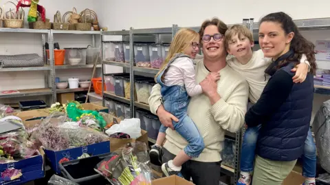 A man is holding a girl and a woman is holding a boy. They all stand inside a shop filled with flowers, shelves and transparent boxes. The girl on the left has long red hair, glasses, a pink fluffy cardigan, a pink t-shirt and denim dungarees. She is being held by a man with long fair hair, glasses, a white woolly jumper and black trousers. The boy has short fair hair, a long sleeved white top and has an arm around the man. His other arm is around the woman who is carrying him. She has long dark hair, green jeans and a navy bodywarmer covering a black top. Behind them are rows of shelves with labelled transparent boxes full of different coloured flower petals. In front of them are trestle tables covered in bunches and boxes of flowers. Other shelves at the back of the shop have baskets stacked on them.
