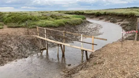 A wooden makeshift bridge at Stiffkey marshes. There is a channel of water running underneath with mud and grass on the adjacent banks.