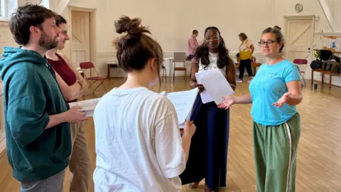 A group rehearsing in a hall. A woman in a blue top is leading the rehearsal.