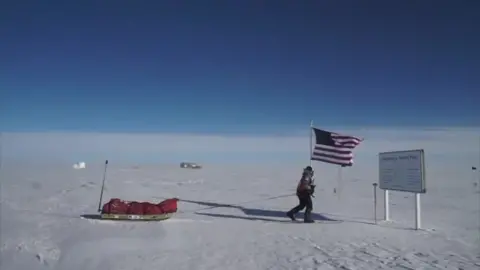 Cat Burford Surrounded by snow Cat Burford skis to a sign making the South Pole. She is dragging a sled with equipment covered by a red material, with the US Stars and Stripes flag just behind her.