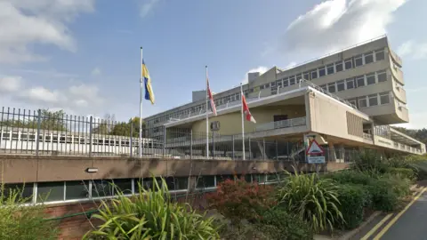 Google Three flagpoles outside a concrete council building. They fly the yellow and blue Ukraine flag, the Union flag and the red and white county flag