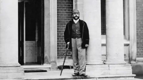Ancient House Museum Maharajah Duleep Singh with walking stick standing beside two columns outside of Elveden Hall. He is wearing a cap, checked trousers dark jacket, and a waistcoat with a watch chain.