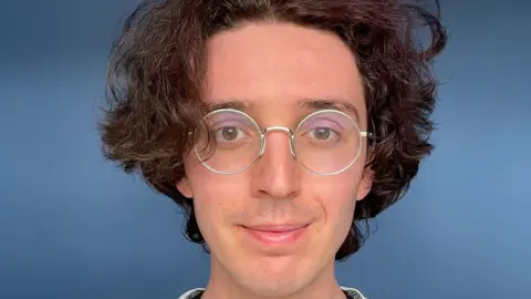 Esme Byrd A picture of a person's face with brown wavy hair and circular glasses in front of a blue background