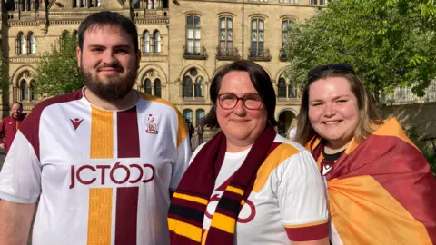 Three Bradford city fans, a man in his 20s, a woman in her 50s and a woman in her 20s wearing Bradford tops outside smiling.