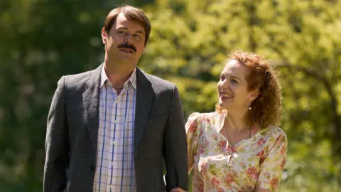 Robert Viglasky/Disney Danny Dyer with a moustache, shirt and grey jacket strolling arm-in-arm with a smiling Katherine Parkinson, wearing a floral dress