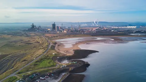 Overhead shot of coastal area, and a large industrial complex with high buildings and chimneys backing onto a large sandy bay, with further industrial buildings to the left. To the right is a scrubby grassed area. Low rising hills can be seen in the background.