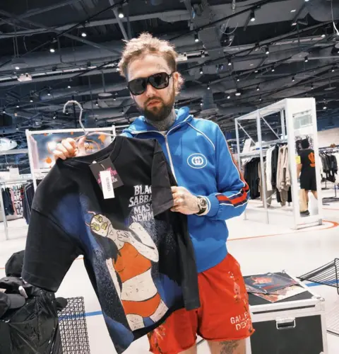 Mason Newman Mason is holding a t-shirt on a hanger and wearing shades, red shorts and a blue zip up tracksuit top.