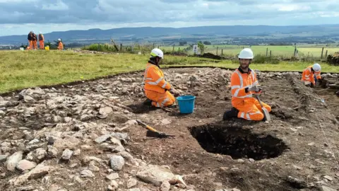 Lots of people in high-vis orange jackets and while helmets digging in red-brown ground with fields and hills in the background.