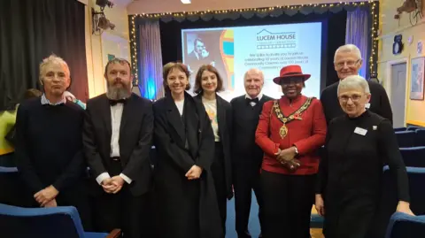 A group of eight people stood in a line smiling at the camera inside Lucem House Community Cinema during its 10 year anniversary