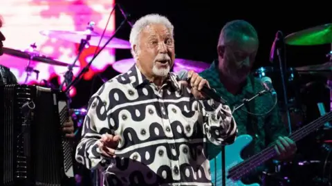 Getty Images Jones has short grey hair and is singing into a mic while wearing a black and white jacket.