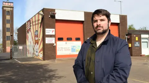 A man with short brown hair and a beard and moustache, wearing a green shirt and black jacket, is standing with a serious expression in front of a small fire station, which has large red doors and a training tower.