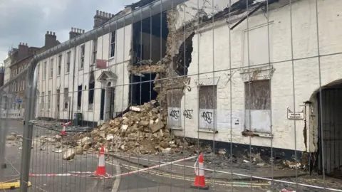 Peter Cooper/BBC Two two-storey white houses side by side. There is a gap in the front facade in which the base of an upper floor can be seen. There is a large pile of rubble including bricks in front of the gap, spilling onto the pavement. The building is fenced off.