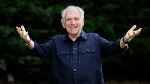 PA Media Hugo Duncan smiling at the camera with his arms outstretched. He is wearing a navy shirt. Behind him there are green bushes.