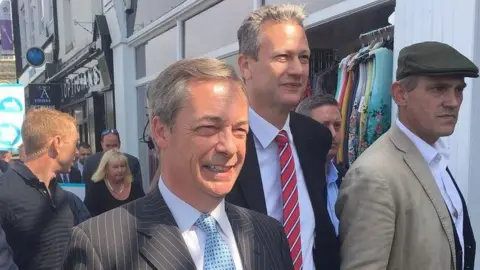 Nigel Farage and Nathan Gill walking at the head of a group of people in a street in Caerphilly. An EU flag can be seen in the background. Farage is at thew centre of the picture, Gill is to his left and slightly behind him. Both men are wearing dark suits, white shirts and ties. 