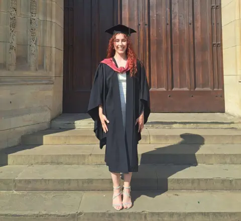 PA Media Caitlin stands outside a university building on the day of her graduation. She is wearing a graduation cap and gown and has long, red hair.