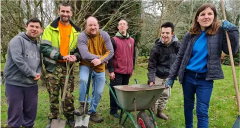 Minstead Trust Five men and a woman standing in a garden holding gardening tools. There is a wheelbarrow next to them