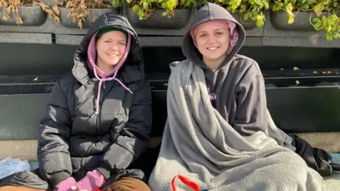 Holly (left) and Ellie (right) sit on the floor in the queue. They wear hoodies with their hoods up. Holly is wearing a black jacket over her jumper. Ellie has a grey blanket pulled up to her left shoulder.