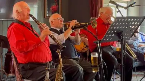 Three members of a jazz band, two wearing red and one in a black waistcoat, play a clarinet, a trumpet and a trombone