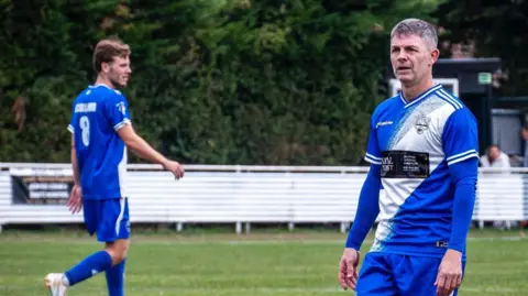 Martell McKenzie Jamie Cureton, wearing a blue and white strip, stands on a football pitch during a match. He is standing, looking off-camera. A team-mate stands behind him. wearing the number 8 shirt. 