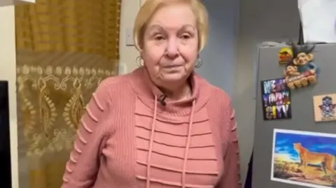 Loraine White, a woman with short blonde hair wearing a pink hoodie, stands in the doorway to her kitchen. A grey fridge with colourful magnets can be seen to the right of the image