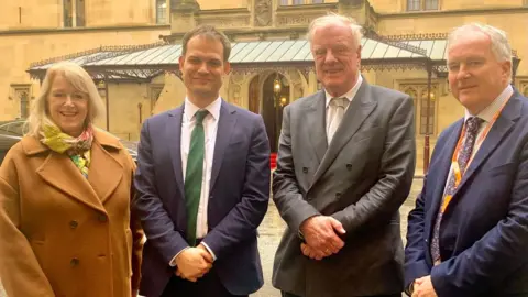 Local Democracy Reporting Service West Lindsey District Council deputy leader Lesley Rollings, Lincoln MP Hamish Falconer, Gainsborough MP Sir Edward Leigh, and council leader Trevor Young smiling and facing the camera outside a building at Westminster. 