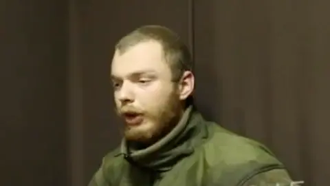 BBC A man believed to be James Scott Rhys Anderson, 22, a former British Army soldier captured while fighting for Ukraine, is seen wearing green military clothing and sat in front of a plain brown background.